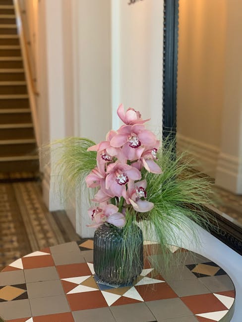 A tall floral arrangement featuring multiple pale pink orchids with delicate petals and central white and purple markings, arranged in a dark, ribbed glass vase. The orchids are complemented by wispy