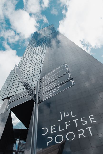 A close-up view of a modern glass office building reaching into a partly cloudy sky, featuring reflective surfaces that mirror the surrounding clouds and blue sky. Prominently displayed on the buildin