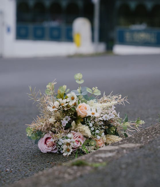 A delicate bouquet of fresh flowers lies on the pavement near the edge of a road, featuring soft pink roses, small white daisies, and pale peach peonies, complemented by muted green eucalyptus leaves