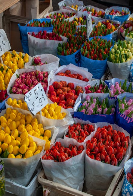 A vibrant display of numerous flower bouquets featuring tulips arranged in large paper-wrapped bundles. The tulips come in a variety of colors, including bright yellow, deep pink, orange, red, purple,