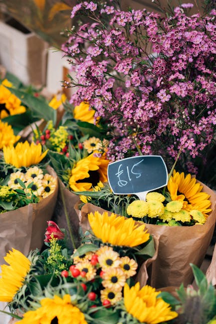 A vibrant arrangement of fresh flowers including bright yellow sunflowers, small white daisies with yellow centers, and red and yellow blossoms wrapped in brown paper, displayed outdoors on a market s
